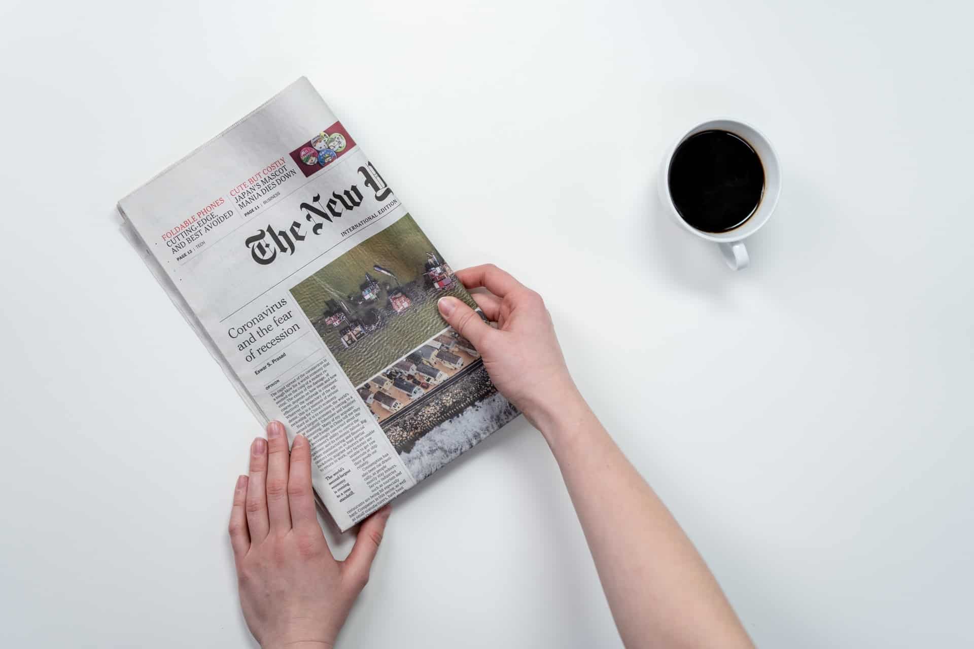 hands holding a folded newspaper with a cup coffee next to it